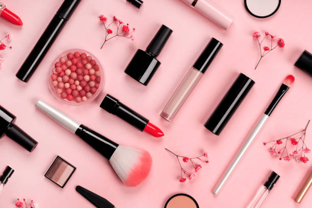 トレンディなパステル ピンクの背景に赤い花とメイクやマニキュア用のさまざまな化粧品アクセサリー。チーク、ブラシ、アイシャドウ、マスカラ、香水、口紅、マニキュア。スキンケア商品。
