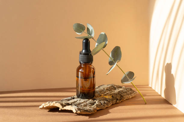 Flaska eterisk eukalyptusolja i en bärnstensfärgad flaska med dropplock på brun bakgrund, i solljus.Kvist med gröna blad på stativ av trädbark.Begreppet aromaterapi.