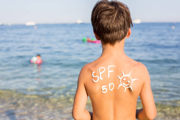 Fëmijë, djalë para adoleshent me krem ​​mbrojtës nga dielli në shpinë në plazh, duke mbajtur unazë të fryrë