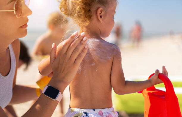 Caring mother apply sunblock to the back of her little daughter. Summer vacation sea beach. Caucasian family with one child resting. Lifestyle photo. Sun protection cream.